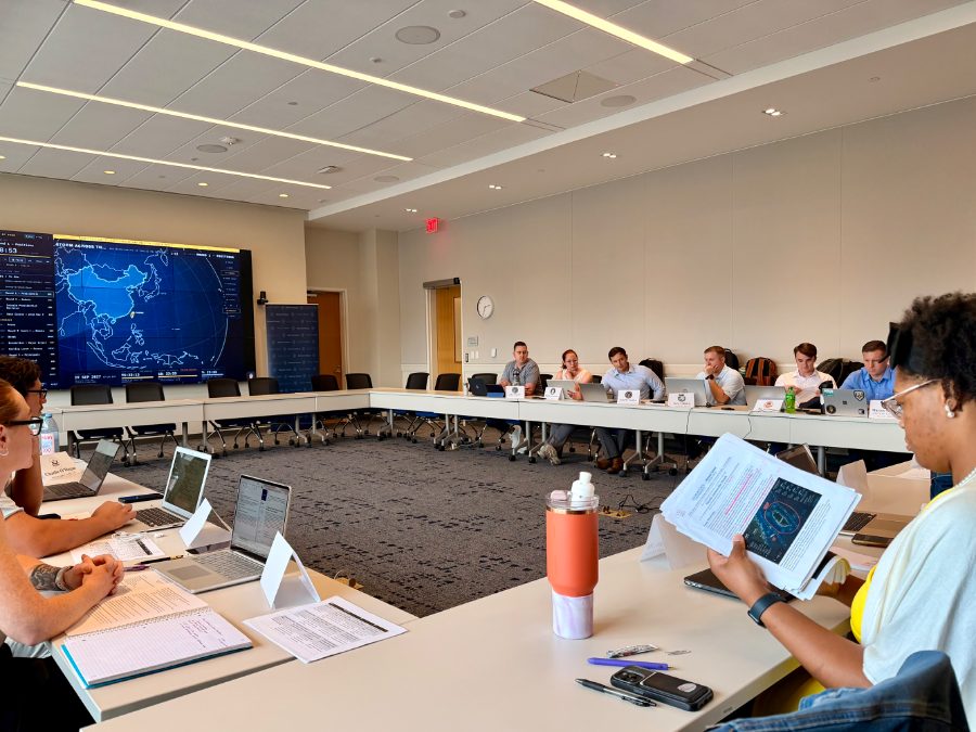 A seminar room mid-exercise: participants at a hollow-square table with named seat placards, the run-of-show and a Western Pacific map on the wall screen.